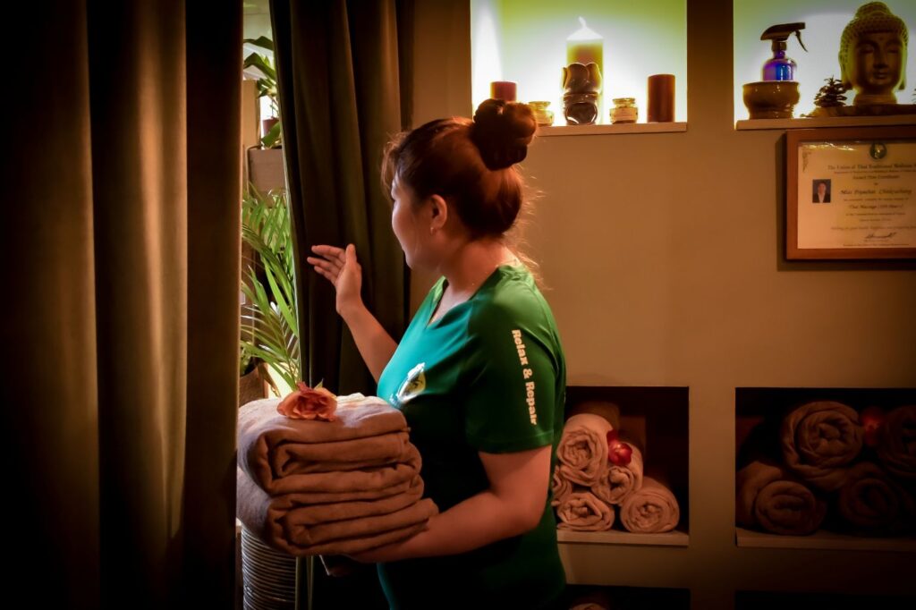 Bonzai24 massage-therapeut in groene spa-kleding met opgestoken haar opent een gordijn naast een raam en houdt opgevouwen handdoeken vast in een rustgevende spa-ruimte met Buddha-beeld, kaarsen en opgerolde handdoeken.