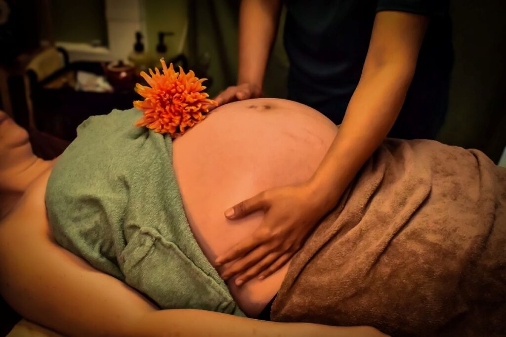 Zwangere vrouw ontvangt een ontspannende zwangerschapsmassage op haar buik in een warme spa-omgeving, met handdoeken en een bloem als decoratie.