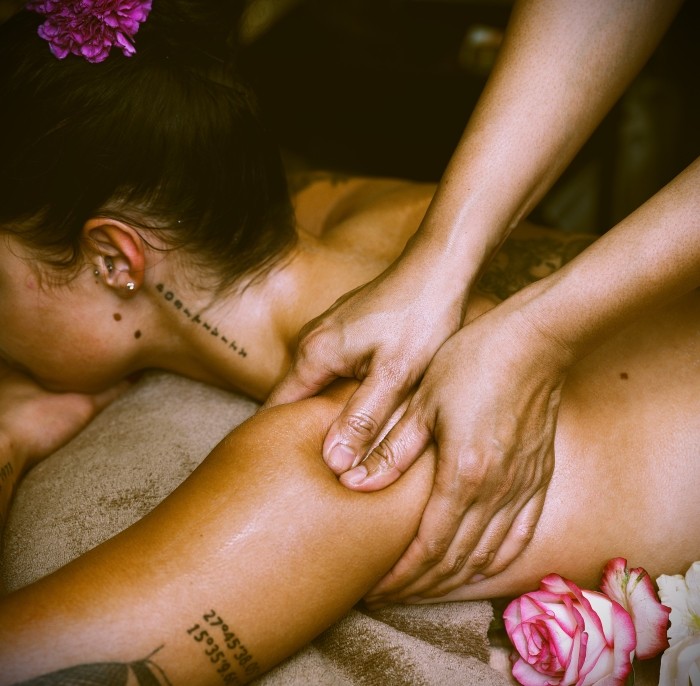 Close-up van een helende massage waarbij een persoon stevig op de schouder van een vrouw drukt. De vrouw ligt ontspannen op haar buik met bloemen in haar haar, en de sfeer is warm en rustgevend. Tatoeages op haar arm zijn zichtbaar.