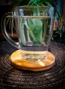 Glazen mok met helder water op een ronde houten onderzetter, geplaatst op een donker geweven oppervlak met groene kamerplanten op de achtergrond.
