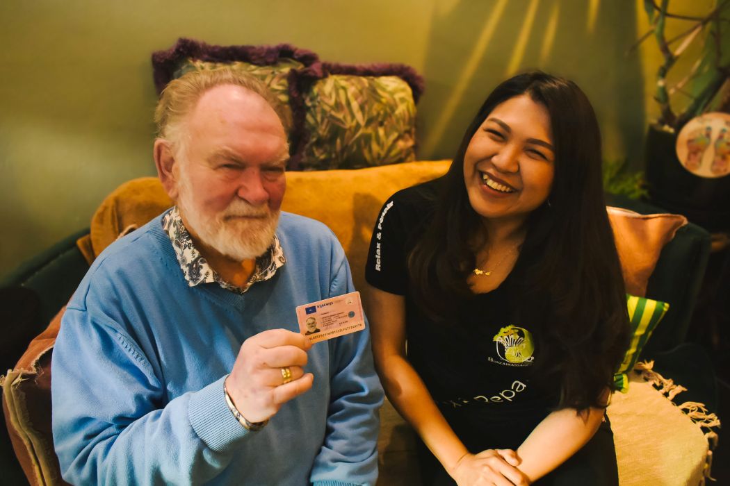 Een oudere man met een witte baard zit op een bank en houdt een identiteitskaart omhoog. Naast hem zit een jonge vrouw die glimlacht naar de camera. Beiden lijken ontspannen in een gezellige huiselijke omgeving.