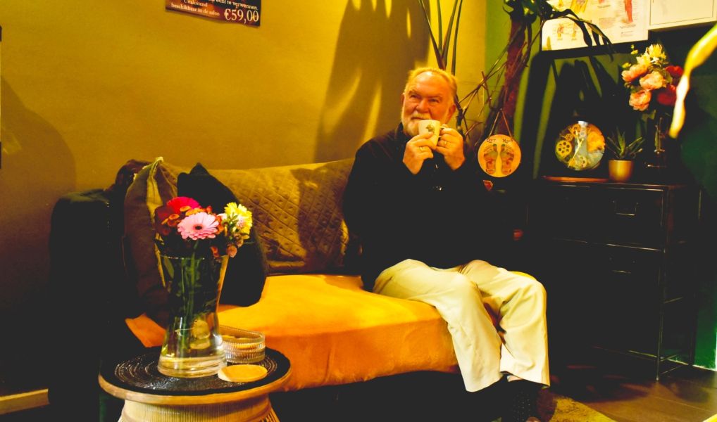 Oudere man ontspant op een bank bij Bonzai24 met een kop koffie in een warme, sfeervolle woonkamer met bloemen, planten en decoratieve accessoires.