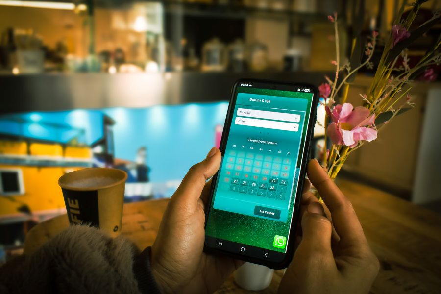 Persoon gebruikt een smartphone in een café in Amersfoort om online een datum en tijd te reserveren via een reserveringsapp, met koffie en bloemen op tafel.